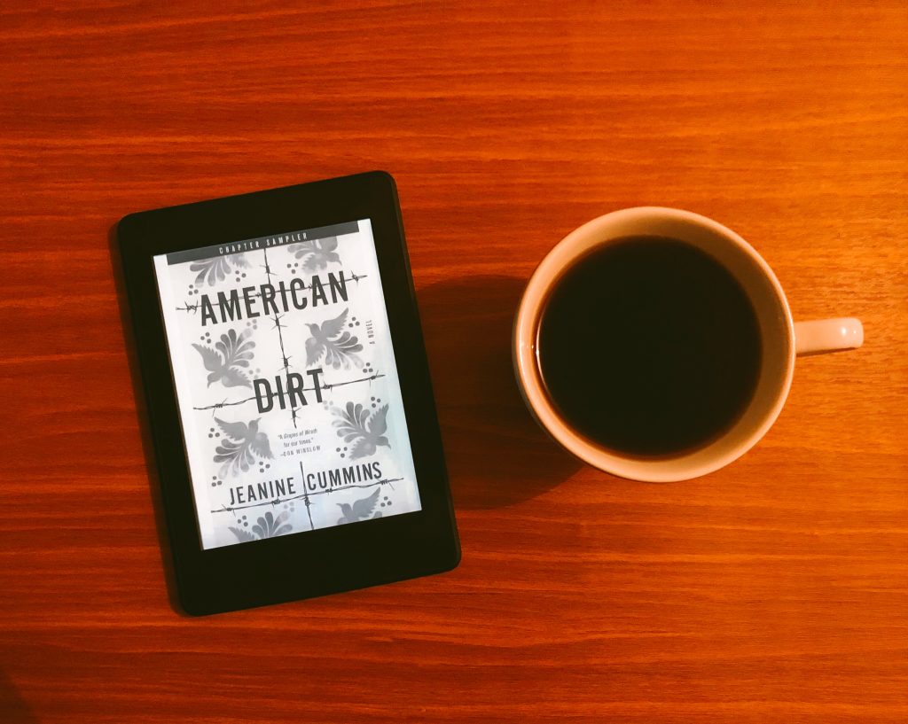 A foto mostra um kindle exibindo a capa do romance American Dirt de Jeanine Cummins à esquerda e uma xícara de chá à direita. Ao fundo, uma mesa de madeira.