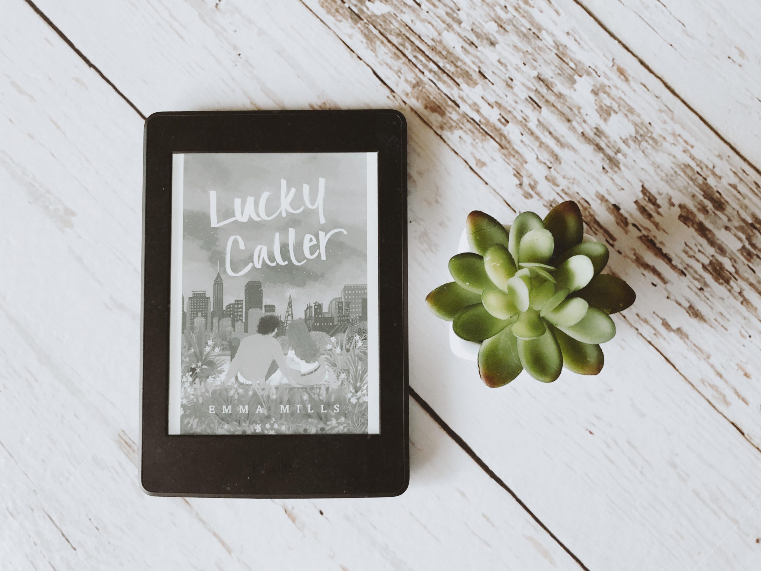 Foto de um Kindle exibindo a capa de Lucky Caller de Emma Mills à esquerda e uma planta suculenta à direita. Ao fundo, uma textura de madeira branca.