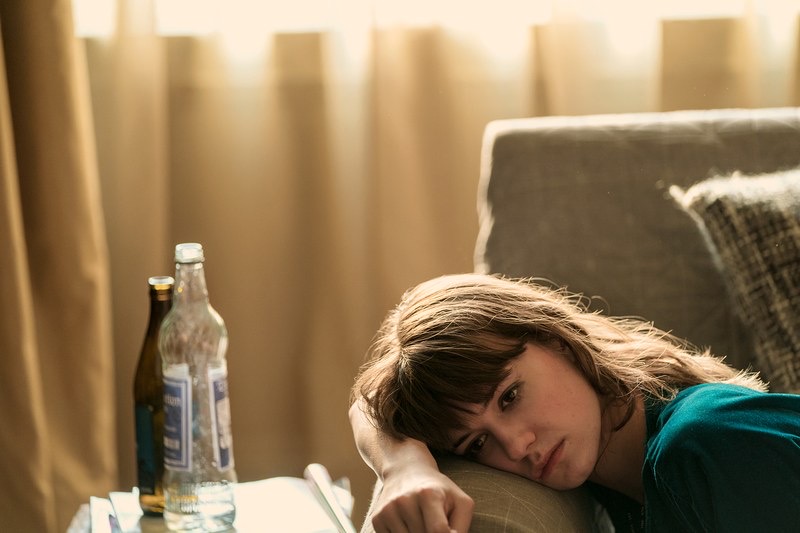 Daisy Edgar-Jones como Marianne do romance NORMAL PEOPLE de Sally Rooney. Daisy está deitada com a cabeça no braço do sofá com ar triste. À direita, em uma mesa, duas garrafas.
