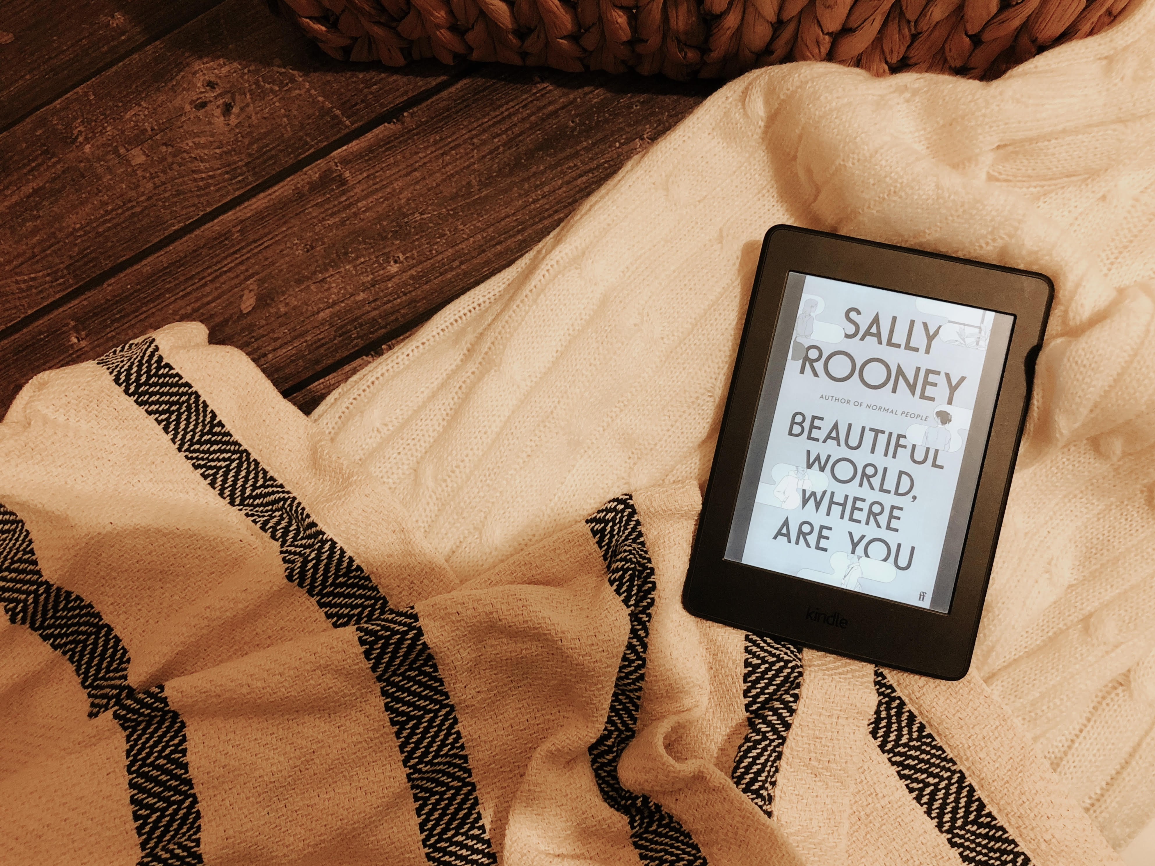 Foto de um Kindle paperwhite ligado mostrando a capa do romance "Beautiful World, Where Are You" sobre uma manta branca à direita e uma manta com listras brancas e pretas à direita.