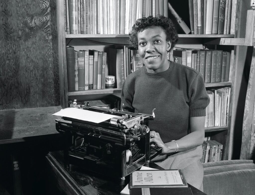 A imagem mostra a autora, Gwendolyn Brooks, com uma máquina de escrever e livros ao fundo, em preto e branco.