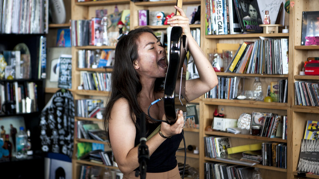 A imagem apresenta a cantora Mitski segurando uma guitarra na altura da cabeça, gritando em direção as cordas em uma performance de "Class of 2013".