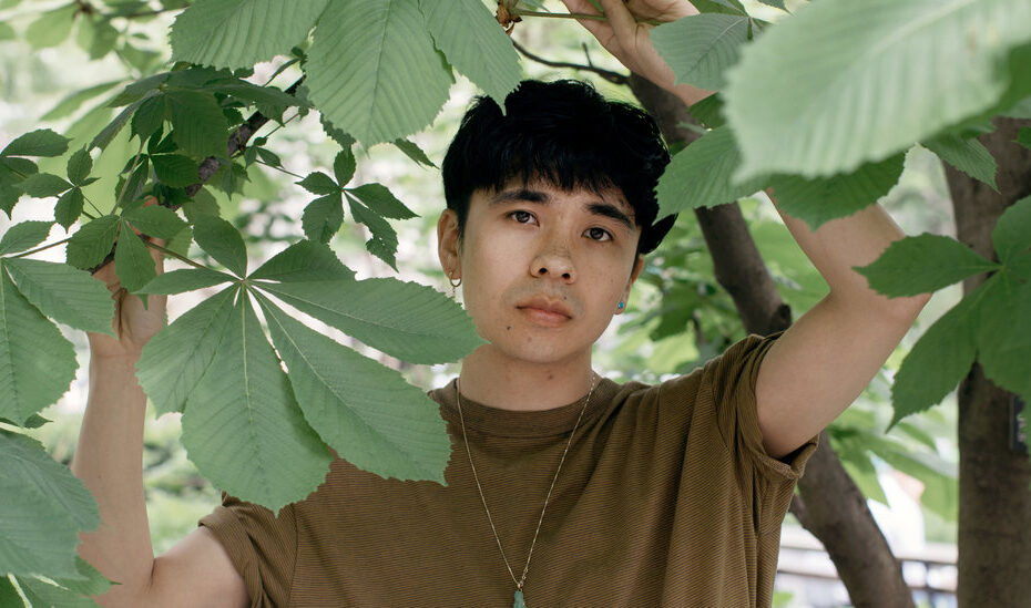 A imagem colorida apresenta o autor ocean Vuong em meio a algumas plantas. A foto é colorida e o homem tem cabelos escuros e utiliza blusa marrom.