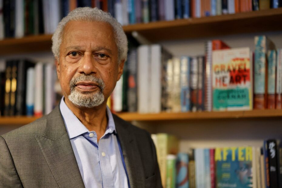 A imagem apresenta uma foto do autor Abdulrazak Gurnah, um homem negro vestido de terno, com cabelo e barba grisalhos.