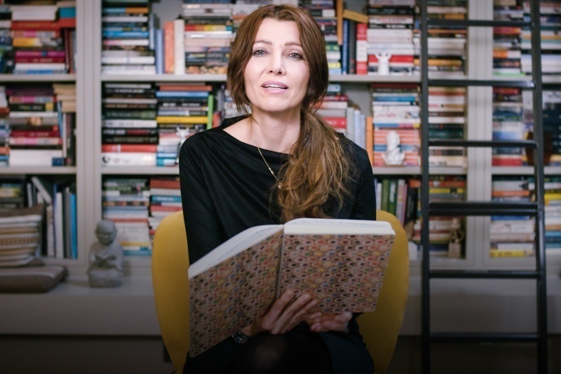 A imagem mostra a autora Elif Shafak segurando um livro em uma biblioteca