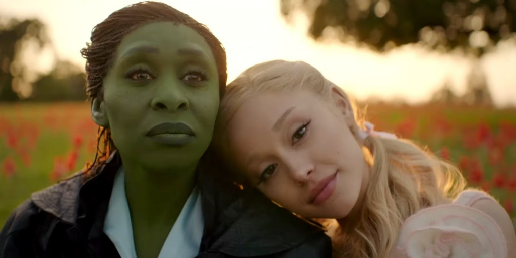 A imagem é um trecho do filme "Wicked", em que Galinda (Ariana Grande) aparece apoiada no ombro de sua amiga, Elphaba (Cynthia Erivo)